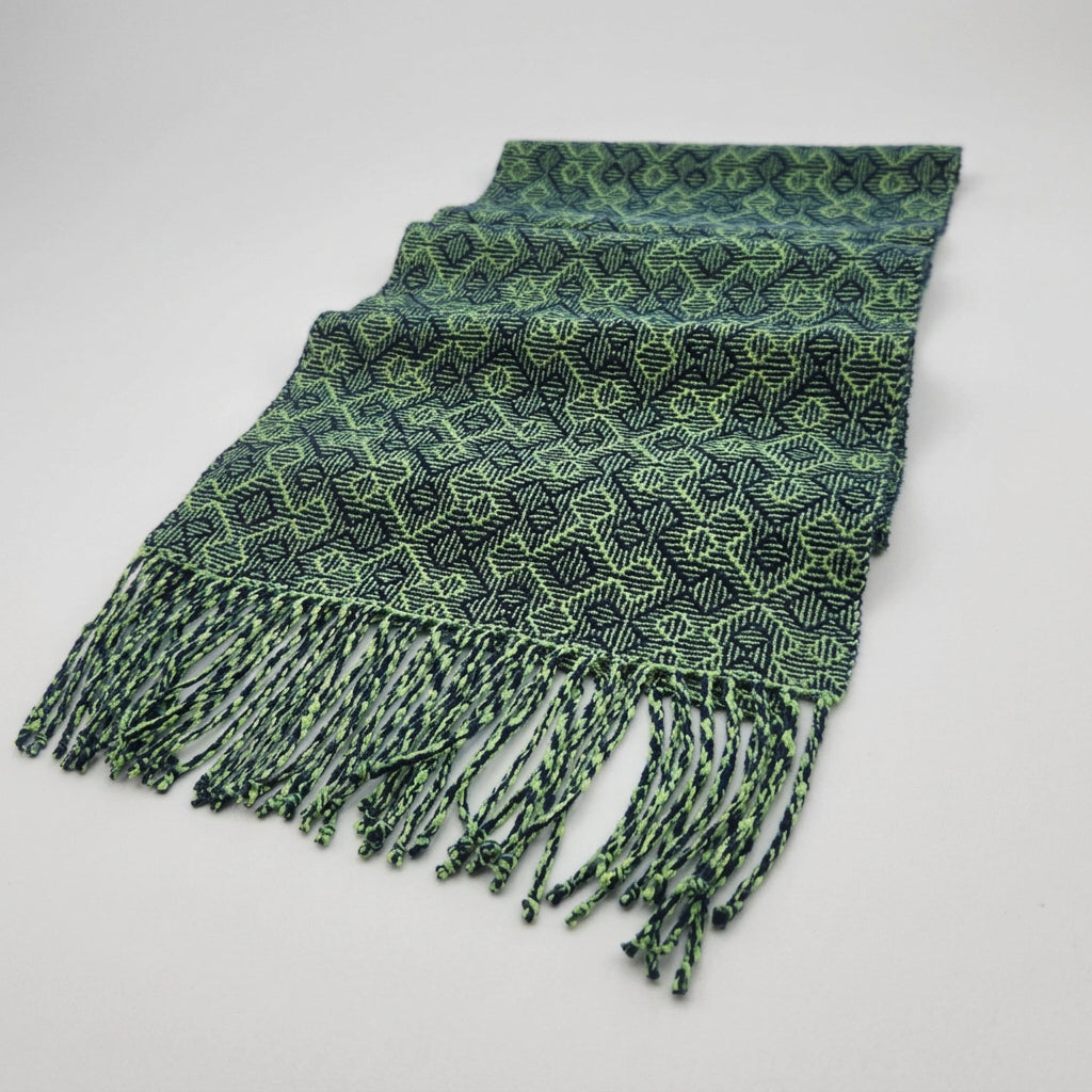 A green and black patterned woven scarf with fringe, laid out on a white table in front of a window showing a garden view.